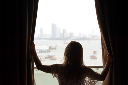 Asian woman opens brown window curtains with boats and building in the bay background.