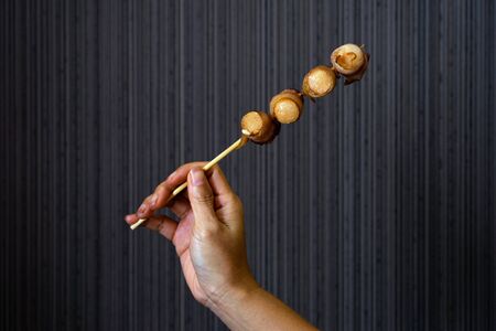 Asian female hand holding bacon wrapped sausage skewer on natural light and dark background.
