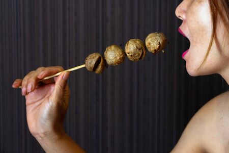 Asian female with pink lips holding and eating meatballs skewer on dark background.の写真素材