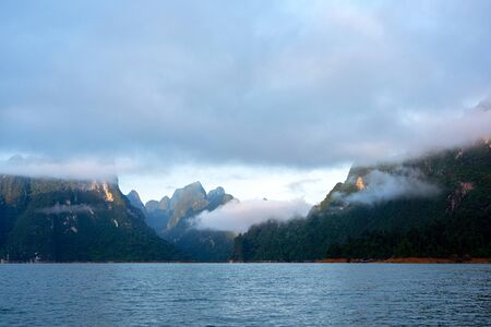 Clouds and mist over green limestone mountain range in the morning light with blue lake in Khao Sok...