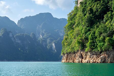 Green trees on rocky cliff with limestone mountain range, lake and blue sky background in Khao Sok...