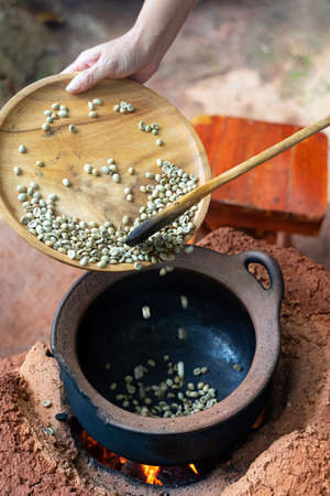 Female hand holding wooden tray pouring raw coffee bean into black clay pot on fire stove.の写真素材