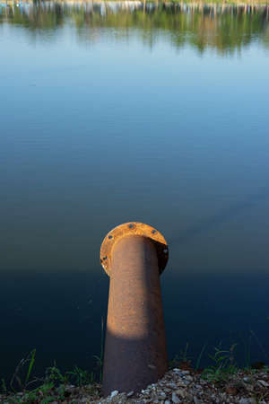 Old rusty steel drainage pipe under sunlight with blue water and green reflection of trees.