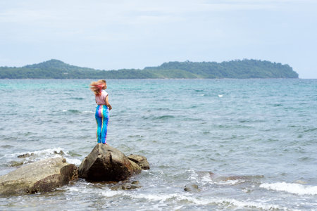 Asian woman standing on a large stone with wind blown hair at the sea on Koh Kood island, Thailand.の写真素材