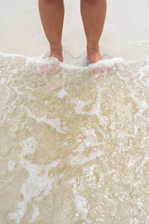 Asian female legs standing on sand beach with small waves touching her feet.の写真素材