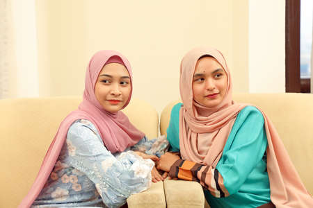 Two young Asian Malay Muslim woman wearing headscarf at home office student sitting on sofa talk...