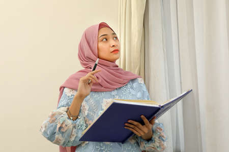 young Asian Malay Muslim woman wearing headscarf at home office next to window look at book document study think writeの写真素材