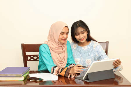 Two young Asian Malay Muslim woman wearing headscarf at home office student sitting at table talk mingle look at computer book document study discuss read type point showの写真素材