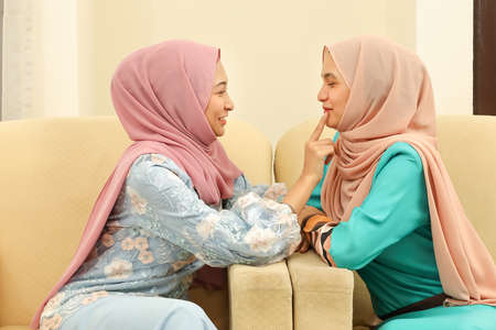 Two young Asian Malay Muslim woman wearing headscarf at home office student sitting on sofa talk mingle look at each other smile happyの写真素材