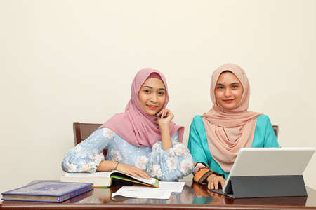 Two young Asian Malay Muslim woman wearing headscarf at home office student sitting at table talk...