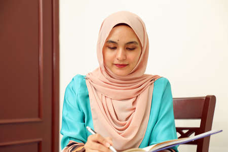 young Asian Malay Muslim woman wearing headscarf at home office student sitting at table read write book document note study smile happy lookの写真素材