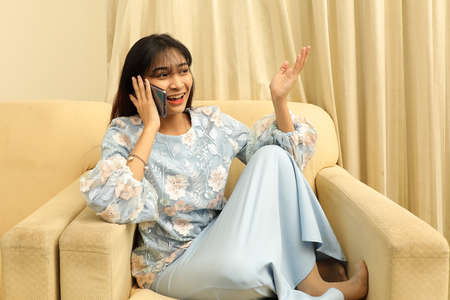 young Asian Malay Muslim woman wearing baju kurung dress at home sit rest on sofa with feets up talk...