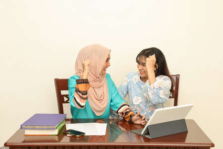 Two young Asian Malay Muslim woman wearing headscarf at home office student sitting at table talk...