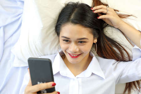 Young beautiful modern Malay woman lying on white pillow bed wearing white shirt using smartphone...