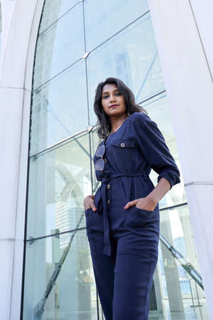 Young attractive beautiful Indian woman in dark blue formal jumpsuit look forward confidently hands in pocket front of a tall glass urban buildingの写真素材
