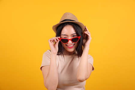 Beautiful young south east Asian woman red frame sunglass hat pose style fashion peak happy on yellow orange background hand behind head pull downの写真素材