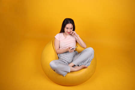 Beautiful young south east Asian woman sits on a yellow beanbag seat orange yellow color background pose fashion style elegant beauty mood expression rest relax think emotion sleepの写真素材