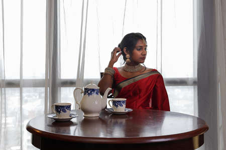 Indian woman wearing red orange traditional royal saree and jewellery siting vintage table tea pot cup bright window backlightの写真素材