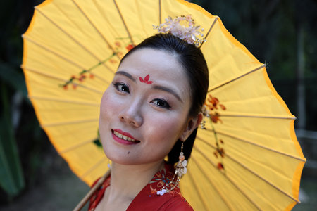Young attractive Asian woman wearing tadeonal Chinese red hanfu long skirt dress costume scarf hairpin earing decorated umbrella outdoor green garden patioの写真素材
