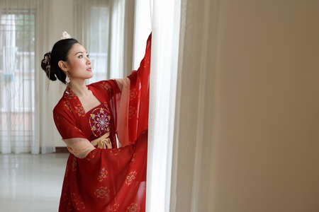 Young attractive Asian woman wearing tadeonal Chinese red hanfu long skirt dress costume scarf hairpin earring indoor at window white curtain lightの写真素材