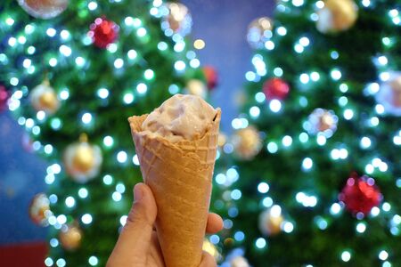 In selective focus a female hand holding a cone of melting vanilla ice cream with beautiful bokeh light Christmas tree at nightの写真素材