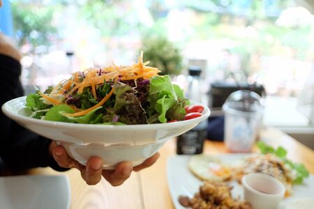 A white plate of fresh vegetable salad on a female hand holding with blurred a dinning table area in the restaurantの写真素材