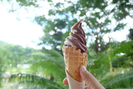 In selective focus a female hand holding a cone of chocolate ice cream with blurred green bokeh and nature background in a garden areaの写真素材