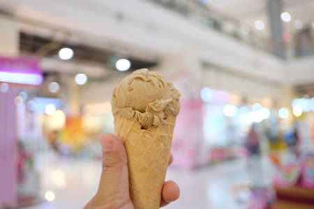 In selective focus a female hand holding a soft cone of coffee ice cream with blurred the mall area...