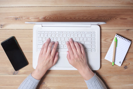 Top view of a wooden table with a laptop in the center. A woman's hands are visible, typing on the...