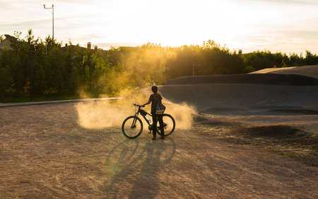 Kazakhstan, Nur-Sultan, July 2021. A teen cyclist stands in a cloud of dust. The photo was taken in the park at sunset.の写真素材