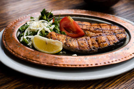 Grilled salmon steak. Served on a tin plate with arugula, lemon, tomato and feta cheese.