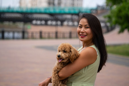 A beautiful Asian girl (Kazakh) holds a dog (mini poodle) in her hands. The girl has emotions of...