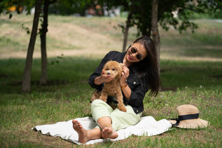 A beautiful Asian girl (Kazakh woman) feeds her dog (mini poodle) from her hands. Summer portrait of a young woman in the park. high quality photoの写真素材