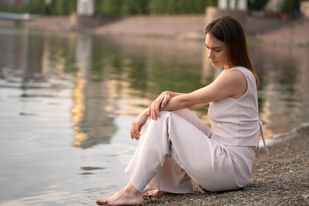 Summer urban portrait of a young brunette woman. The woman sits on the bank of the river and thinks about something. high quality photoの写真素材