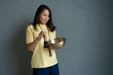 A young Asian woman holds a Tibetan singing bowl in her hands on a gray background with space for text. high quality photoの写真素材
