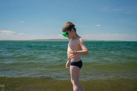 A 7-year-old boy in a diving mask stands on the beach near the water.の写真素材