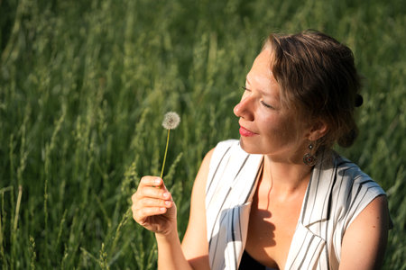 A young blonde woman sits on the grass and blows on a dandelion. high quality photoの写真素材