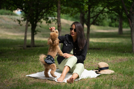 A beautiful Asian girl (Kazakh woman) feeds her dog (mini poodle) from her hands. Summer portrait of a young woman in the park. high quality photoの写真素材