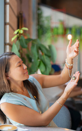 A young cheerful Asian woman sits at a table in a cafe and holds a necklace with a pendant in her hands. high quality photoの写真素材