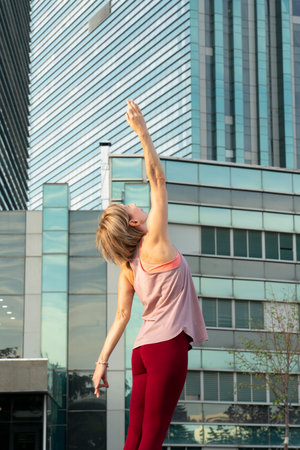 Portrait of a mature woman, 59 years old, doing yoga on a city street. Behind the woman is a blue skyscraper. Active sport lifestyle of mature people.の写真素材