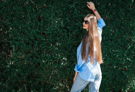 Summer portrait of a girl with long blond hair in sunglasses. A girl in a blue shirt and blue jeans stands against a background of greenery. high quality photoの写真素材