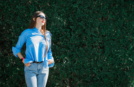 Summer portrait of a girl with long blond hair in sunglasses. A girl in a blue shirt and blue jeans stands against a background of greenery. high quality photoの写真素材