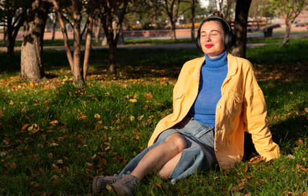 A young woman sits on the green grass in the park and enjoys music on her headphones. High quality photoの写真素材