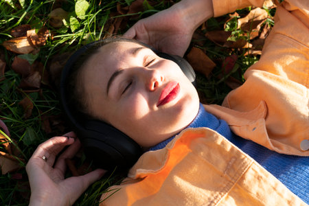 A young woman lies on the green grass in the park and enjoys music on her headphones. High quality photoの写真素材