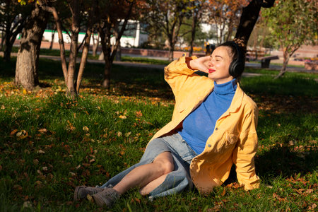 A young woman sits on the green grass in the park and enjoys music on her headphones. High quality photoの写真素材