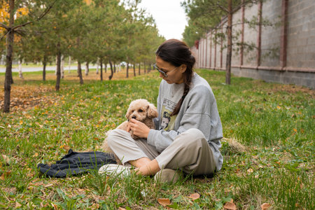 A young Asian woman sits on the grass and feeds her toy poodle. Autumn landscape. High quality photoの写真素材