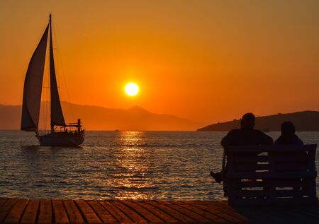Sailing boat on the sea at sunset. Old couple sitting on a bench near the sea.の写真素材