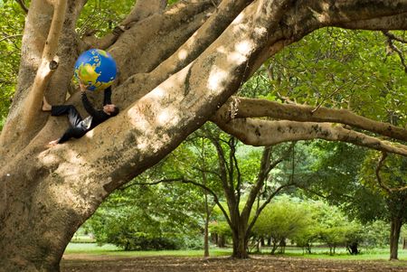 Latin american businessman holding a globe in his hands while sitting in the crook of a tree trunkの写真素材
