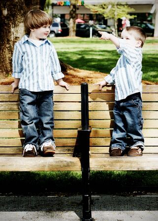 Two young identically dressed brothers standing on a park bench. Vertically framed, high-contrast shotの写真素材