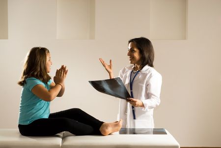 Female Doctor holds an x-ray and smiles as she speaks to a young girl.  The barefoot girl sits on the examining table. Both people look happy with the results as the girl claps her hands in delight. Horizontally framed photographの写真素材